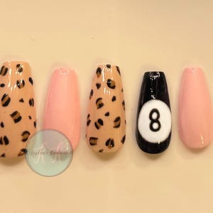 May include: A set of five press-on nails with a glossy finish. Two nails feature a tan base with a black and brown leopard print pattern. Two nails are solid light pink. One nail is black with a white circular accent containing the number 8.
