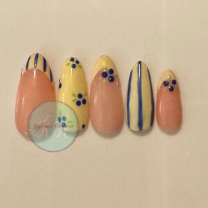 May include: Five almond-shaped press-on nails featuring a mix of designs. Two nails have vertical blue stripes on a cream base, two have a nude base with blue berry accents, and one is a solid cream color with small blue berry illustrations.
