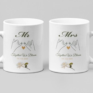 Pode incluir: Duas canecas de cerâmica branca com alças, cada uma com um design com as palavras "Mr" e "Mrs" em escrita elegante. O design inclui mãos entrelaçadas, um coração dourado e o texto "Together We Bloom" com uma data. Uma ilustração floral está na parte inferior.