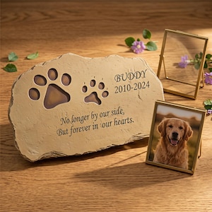 May include: A tan stone pet memorial marker featuring two engraved paw prints, the name BUDDY, the years 2010-2024, and the text No longer by our side, But forever in our hearts, accompanied by two gold-toned metal picture frames.