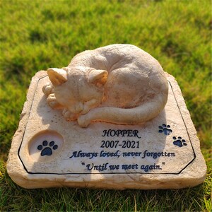 May include: A beige stone memorial with a sleeping cat sculpture. The stone has the inscription "HOPPER 2007-2021 Always loved, never forgotten "Until we meet again."