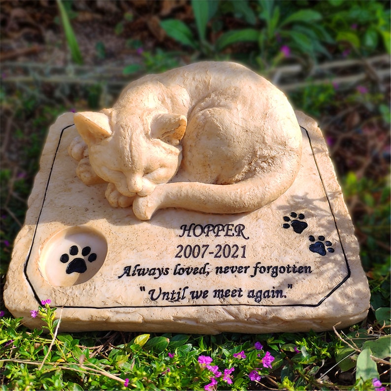 May include: A beige stone memorial with a sleeping cat sculpture. The inscription reads "HOPPER 2007-2021 Always loved, never forgotten "Until we meet again."