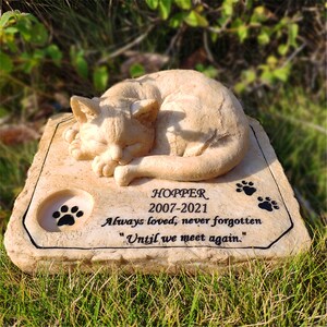 May include: A beige stone memorial with a sleeping cat sculpture. The inscription reads "HOPPER 2007-2021 Always loved, never forgotten "Until we meet again."