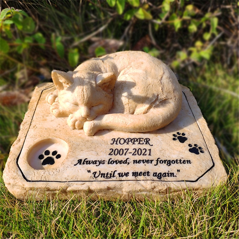 May include: A beige stone memorial with a sleeping cat sculpture. The inscription reads "HOPPER 2007-2021 Always loved, never forgotten "Until we meet again."
