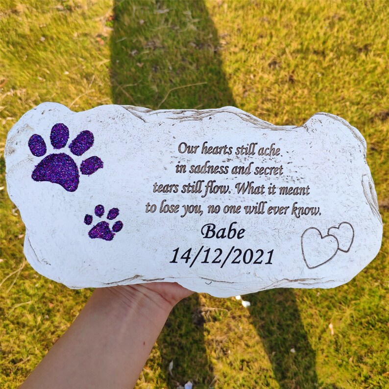 May include: A white stone pet memorial with purple glitter paw prints and engraved text. The text reads "Our hearts still ache... Babe 14/12/2021" with two heart outlines. The stone is held over green grass.