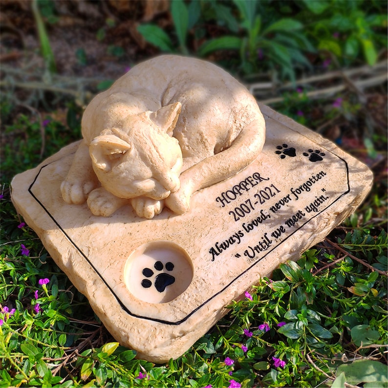 May include: A beige stone memorial with a sleeping cat sculpture. The inscription reads "HOPPER 2007-2021 Always loved, never forgotten "Until we meet again."