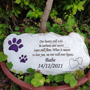 May include: A white stone pet memorial featuring purple glitter paw prints and engraved text. The inscription reads "Our hearts still ache... Babe 14/12/2021". Two hearts are etched on the right. The memorial is set amongst green leaves.