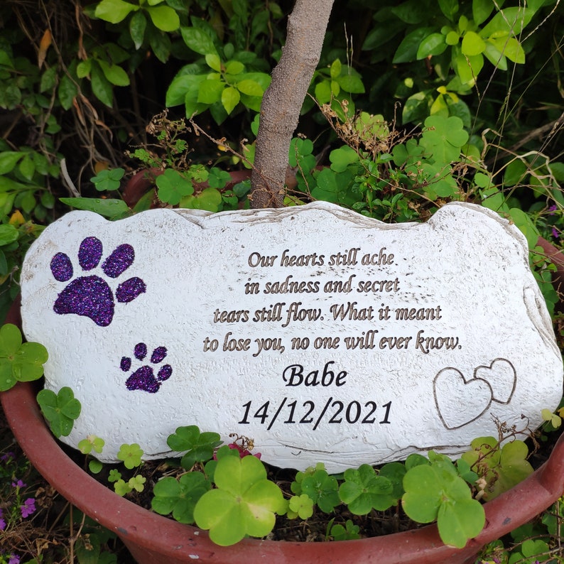 May include: A white stone pet memorial featuring purple glitter paw prints and engraved text. The inscription reads "Our hearts still ache... Babe 14/12/2021". Two hearts are etched on the right. The memorial is set amongst green leaves.