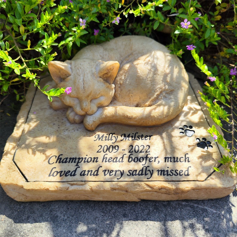 May include: A tan stone memorial plaque featuring a sculpted sleeping cat on top. The text reads: Milly Milster, 2009 - 2022, Champion head boofer, much loved and very sadly missed. Two black paw prints are engraved on the right side of the plaque.
