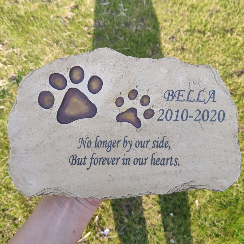 May include: A light tan, irregular-shaped stone memorial marker featuring two engraved paw prints with dark brown centers. The stone is inscribed with the name BELLA, the years 2010-2020, and the text: No longer by our side, But forever in our hearts.