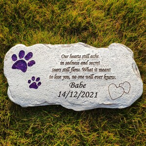 May include: A light grey stone pet memorial with purple glitter paw prints and the inscription "Our hearts still ache... Babe 14/12/2021". Two heart outlines are etched on the right. The stone is set on green grass.