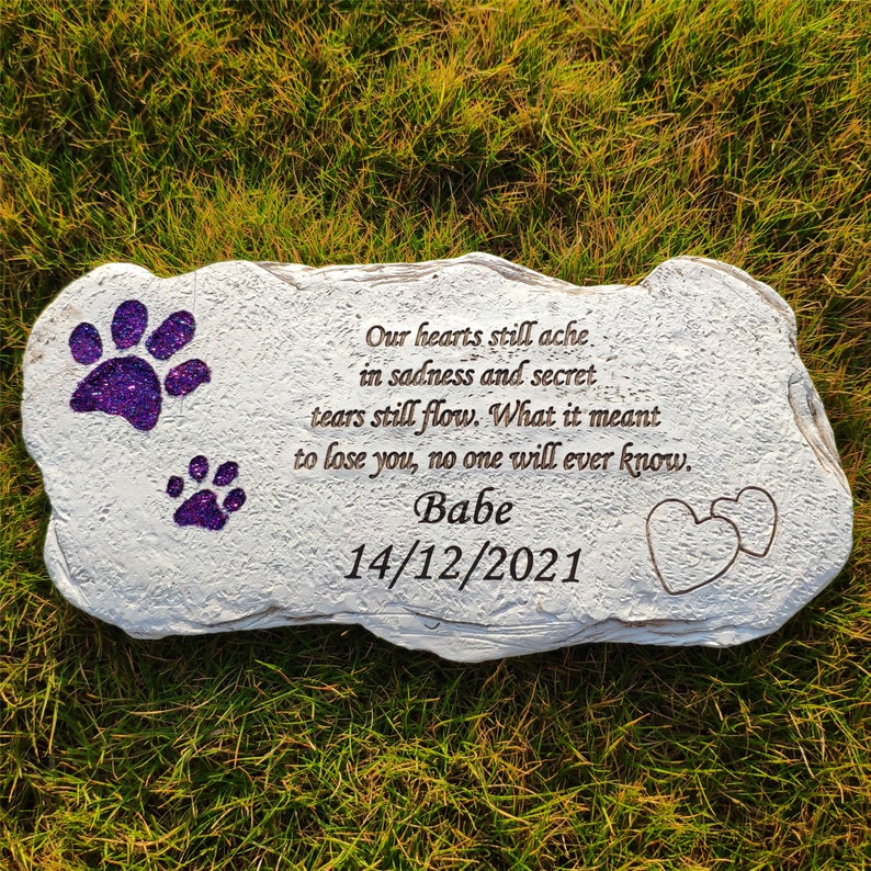 May include: A light grey stone pet memorial with purple glitter paw prints and the inscription "Our hearts still ache... Babe 14/12/2021". Two heart outlines are etched on the right. The stone is set on green grass.