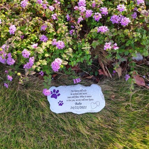May include: A grey stone pet memorial with purple paw prints and the inscription "Our hearts still ache in sadness and secret tears still flow... Babe 14/12/2021." The memorial is set in grass with purple flowers.