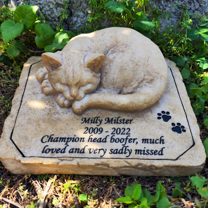 May include: A tan stone pet memorial plaque featuring a sculpted sleeping cat on top. The surface is engraved with the text Milly Milster 2009 - 2022 Champion head boofer, much loved and very sadly missed, accompanied by two black paw print icons.