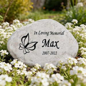 May include: A light gray memorial stone featuring a black butterfly graphic and the engraved text In Loving Memorial, Max, and the years 2007-2022. The stone has a textured, natural surface and is positioned among small white flowers.
