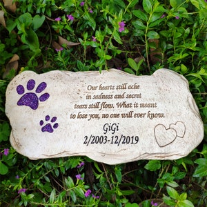 May include: A beige stone pet memorial with purple glitter paw prints and the name "GiGi" with dates. The inscription reads, "Our hearts still ache in sadness and secret tears still flow..." Two hearts are etched on the stone.