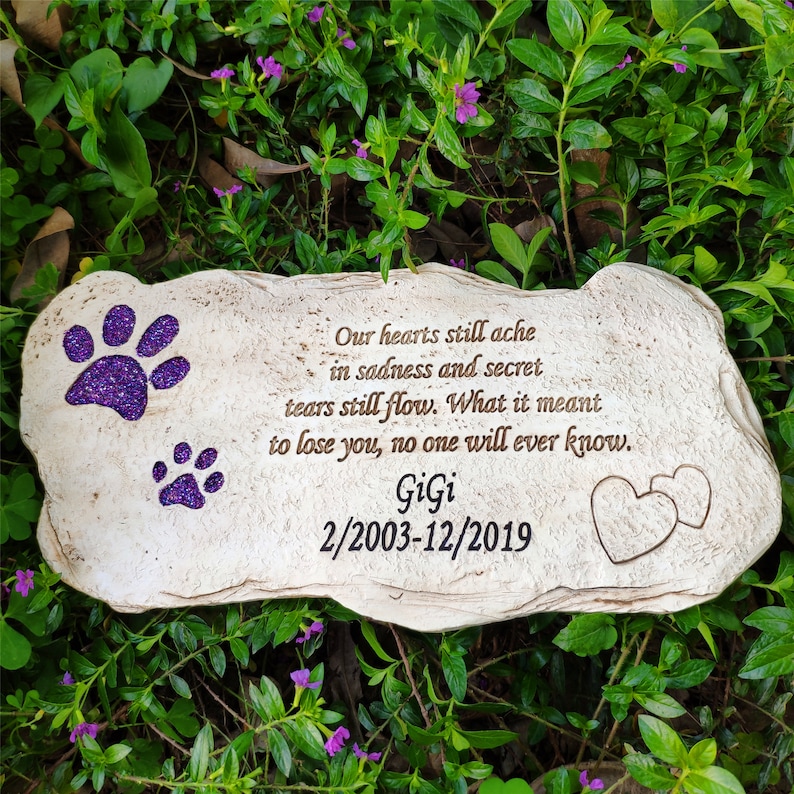 May include: A beige stone pet memorial with purple glitter paw prints and the name "GiGi" with dates. The inscription reads, "Our hearts still ache in sadness and secret tears still flow..." Two hearts are etched on the stone.