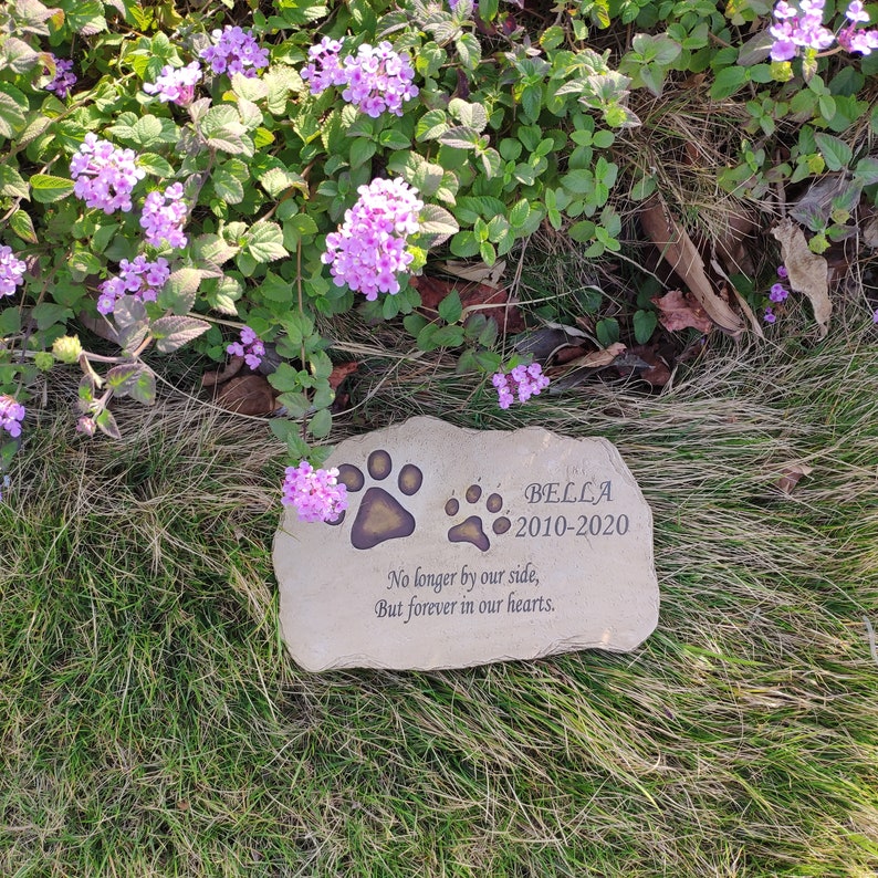 May include: A light tan, rectangular memorial stone with irregular edges featuring two dark brown paw prints, the name BELLA, the years 2010-2020, and the engraved text: No longer by our side, But forever in our hearts. The stone is placed on grass.