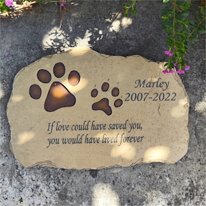 May include: A tan, irregular-shaped stone memorial marker featuring two dark brown paw prints, the name "Marley", the years "2007-2022", and the engraved text "If love could have saved you, you would have lived forever" in a dark serif font.