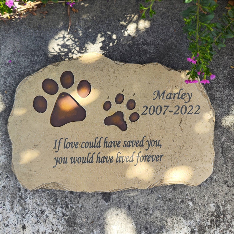 May include: A tan, irregular-shaped stone memorial marker featuring two dark brown paw prints, the name "Marley", the years "2007-2022", and the engraved text "If love could have saved you, you would have lived forever" in a dark serif font.
