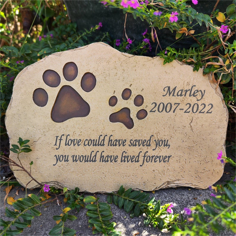 May include: A tan, irregular-shaped stone memorial marker featuring two dark brown paw prints, the name Marley, the years 2007-2022, and the engraved text: If love could have saved you, you would have lived forever, all in a dark, serif font.