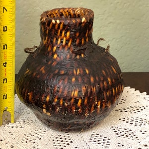 May include: A small, brown woven basket with two handles. The basket is made of natural materials and has a rustic appearance.