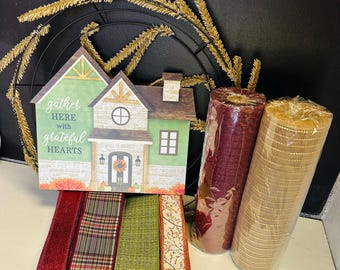 May include: A fall-themed craft kit featuring a house-shaped sign with the text "gather HERE with grateful HEARTS", a black metal wreath frame, rolls of mesh, and various ribbons in fall colors. The ribbons include red glitter, plaid, green, and a floral pattern.