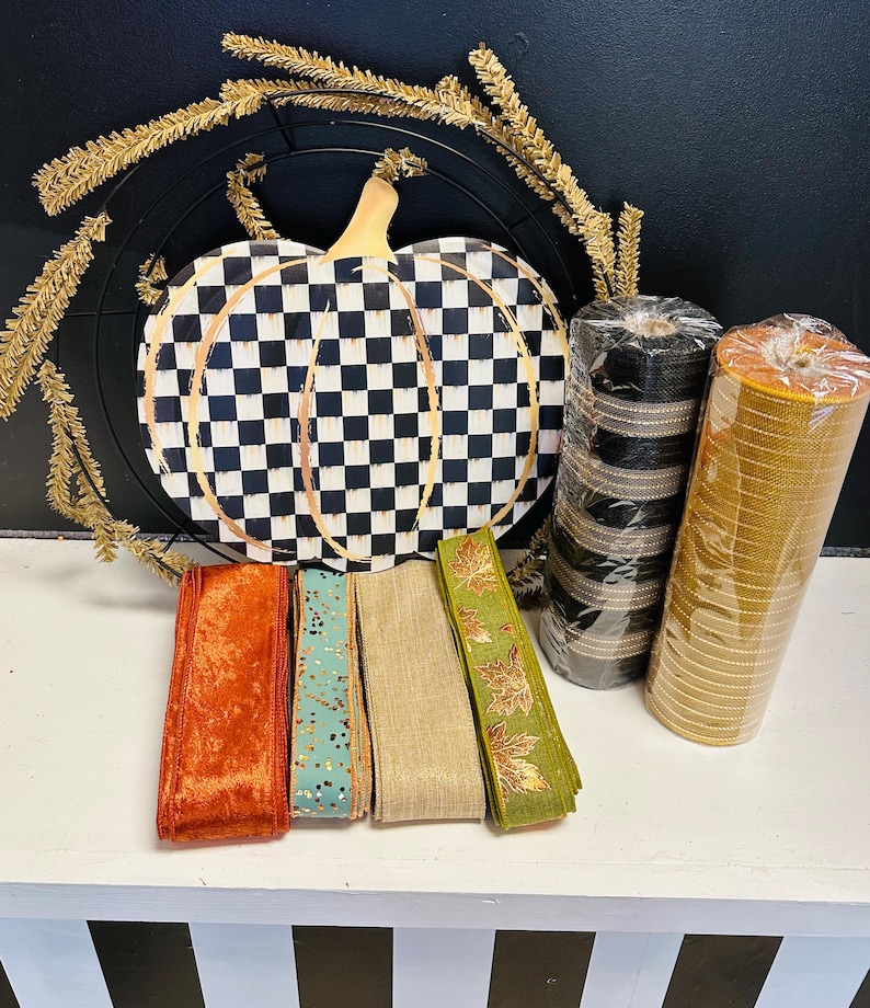 May include: A craft supply set featuring a black and white checkered pumpkin sign, a gold tinsel wreath frame, two rolls of mesh ribbon in black and gold, and four spools of ribbon in orange velvet, teal with gold flecks, tan, and green with gold leaves.