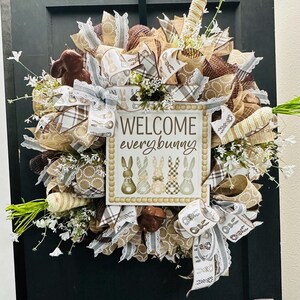 Peut inclure: Couronne de P&acirc;ques avec un panneau "WELCOME every bunny". La couronne pr&eacute;sente des rubans marron, blancs et beiges, de fausses carottes et de petites fleurs blanches. Un lapin en chocolat et des &oelig;ufs d&eacute;coratifs sont &eacute;galement inclus. La couronne est accroch&eacute;e &agrave; une porte noire.
