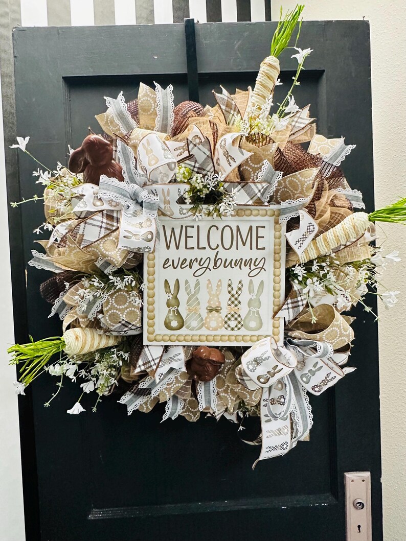 Peut inclure: Couronne de P&acirc;ques avec un panneau "WELCOME every bunny". La couronne pr&eacute;sente des rubans marron, blancs et beiges, de fausses carottes et de petites fleurs blanches. Un lapin en chocolat et des &oelig;ufs d&eacute;coratifs sont &eacute;galement inclus. La couronne est accroch&eacute;e &agrave; une porte noire.