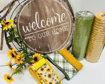 Op de afbeelding: Een rond houten bord met de tekst "Welcome to our home" in witte letters. Het bord is omgeven door een krans van bruine takken en gele zonnebloemen. Er zijn ook verschillende rollen groene en gele lint en een lint met een zonnebloemmotief.