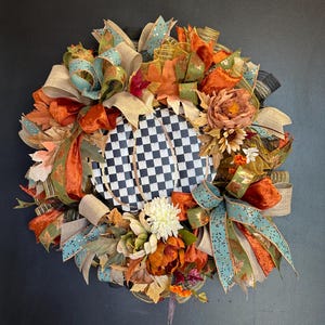 May include: A decorative wreath featuring a central black and white checkered pumpkin shape surrounded by an arrangement of orange, teal, and tan ribbons, artificial autumn leaves, and various flowers in shades of cream, orange, and brown.