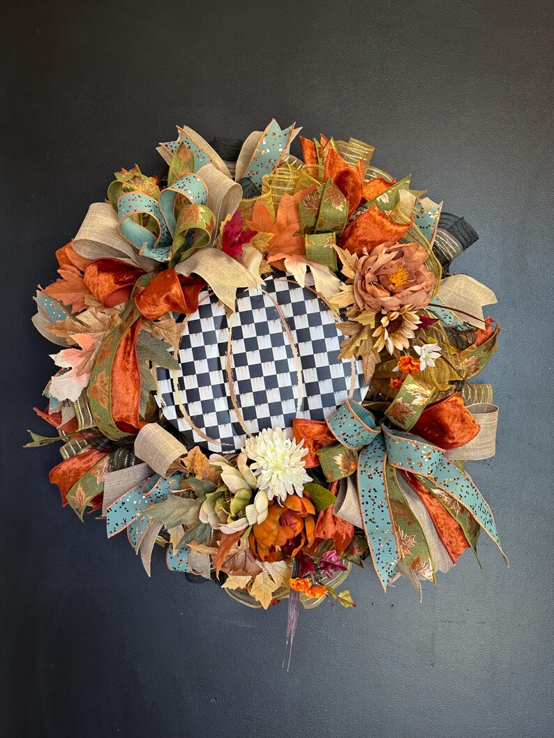 May include: A decorative wreath featuring a central black and white checkered pumpkin shape surrounded by an arrangement of orange, teal, and tan ribbons, artificial autumn leaves, and various flowers in shades of cream, orange, and brown.