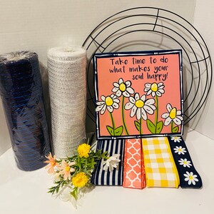 May include: A collection of wreath-making supplies, including rolls of blue and white mesh, decorative ribbons, a metal wreath frame, and a painted sign with daisies and the text "Take time to do what makes your soul happy!"