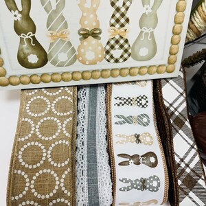 Peut inclure: Une bo&icirc;te d&eacute;corative en bois avec des lapins peints de diff&eacute;rentes couleurs et motifs. En dessous, plusieurs rouleaux de ruban d&eacute;coratif avec diff&eacute;rents motifs, dont des pois, de la dentelle et des motifs de lapins. La palette de couleurs comprend le marron, le blanc et le beige.