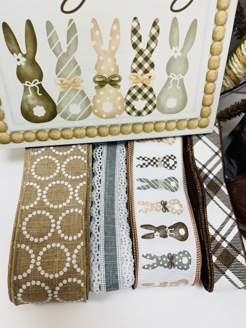 Peut inclure: Une bo&icirc;te d&eacute;corative en bois avec des lapins peints de diff&eacute;rentes couleurs et motifs. En dessous, plusieurs rouleaux de ruban d&eacute;coratif avec diff&eacute;rents motifs, dont des pois, de la dentelle et des motifs de lapins. La palette de couleurs comprend le marron, le blanc et le beige.