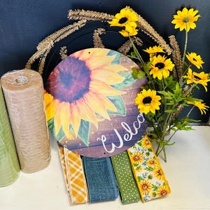 May include: A collection of fall-themed craft supplies. Includes rolls of green and tan mesh, a round wooden sign with a sunflower design and the word "Welcome", and various ribbons with fall patterns. Yellow sunflowers are also present.