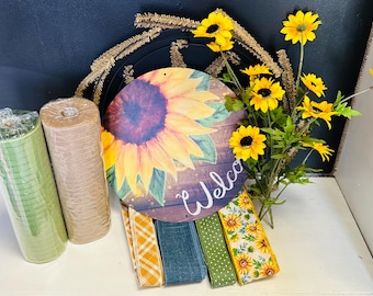 May include: A collection of fall-themed craft supplies. Includes rolls of green and tan mesh, a round wooden sign with a sunflower design and the word "Welcome", and various ribbons with fall patterns. Yellow sunflowers are also present.