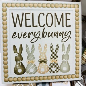 Peut inclure: Panneau carr&eacute; en bois avec l'inscription "WELCOME everybunny" en &eacute;criture marron. En dessous, cinq silhouettes de lapins avec diff&eacute;rents motifs et n&oelig;uds. Le panneau a une bordure perl&eacute;e.