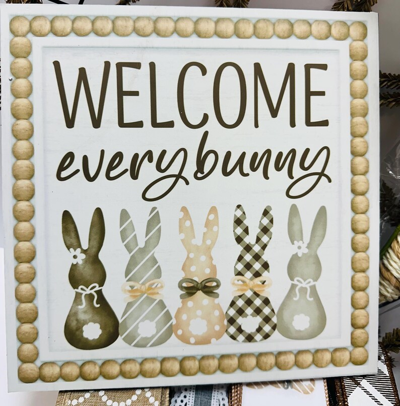 Peut inclure: Panneau carr&eacute; en bois avec l'inscription "WELCOME everybunny" en &eacute;criture marron. En dessous, cinq silhouettes de lapins avec diff&eacute;rents motifs et n&oelig;uds. Le panneau a une bordure perl&eacute;e.