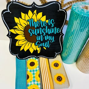 May include: A black wooden sign with a white frame and a sunflower graphic. The sign reads "There is sunshine in my soul". The sign is surrounded by various ribbons and mesh fabric.