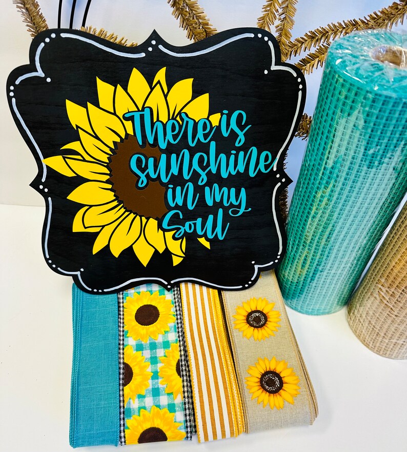 May include: A black wooden sign with a white frame and a sunflower graphic. The sign reads "There is sunshine in my soul". The sign is surrounded by various ribbons and mesh fabric.