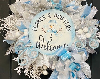 May include: A blue and white winter wreath featuring a snowman and the text "Flakes & Drifters Welcome". The wreath is decorated with white snowflakes, blue and white ribbon, and white mesh.