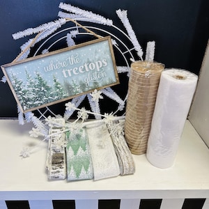 May include: A rectangular wooden sign with the text where the treetops glisten, a white wire wreath frame with white tinsel, four rolls of decorative ribbon in green, white, and brown patterns, and two rolls of mesh fabric in tan and white.