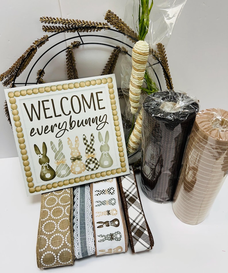 Peut inclure: Un assortiment de fournitures d'artisanat sur le th&egrave;me de P&acirc;ques. Comprend une enseigne avec l'inscription "WELCOME everybunny" avec des illustrations de lapins, des rouleaux de ruban, des carottes d&eacute;coratives et un cadre de couronne en m&eacute;tal. La palette de couleurs comprend du marron, du blanc et du beige.