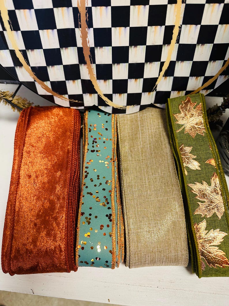 May include: Four rolls of decorative ribbon arranged side by side. The first is a textured burnt orange velvet, the second is a teal ribbon with gold glitter flecks, the third is a solid beige linen-like fabric, and the fourth is olive green with metallic gold maple leaf patterns.