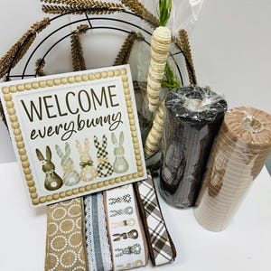 Peut inclure: Un assortiment de fournitures d'artisanat sur le th&egrave;me de P&acirc;ques. Comprend une enseigne avec l'inscription "WELCOME every bunny" avec des illustrations de lapins, des rubans d&eacute;coratifs, des rouleaux de filet et de fausses carottes. Un cadre de couronne en m&eacute;tal est &eacute;galement visible.