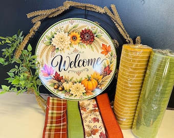 May include: A round metal sign featuring the word Welcome in black script, surrounded by sunflowers, dahlias, pumpkins, and autumn leaves. The sign is set on a black wire wreath frame with two rolls of mesh ribbon and four strips of decorative fabric ribbon.