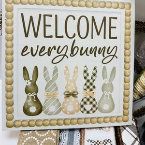 Peut inclure: Panneau carr&eacute; en bois avec l'inscription "WELCOME everybunny" en lettres marron. Le panneau pr&eacute;sente cinq silhouettes de lapins avec diff&eacute;rents motifs et n&oelig;uds. La bordure est faite de petites perles de bois rondes.
