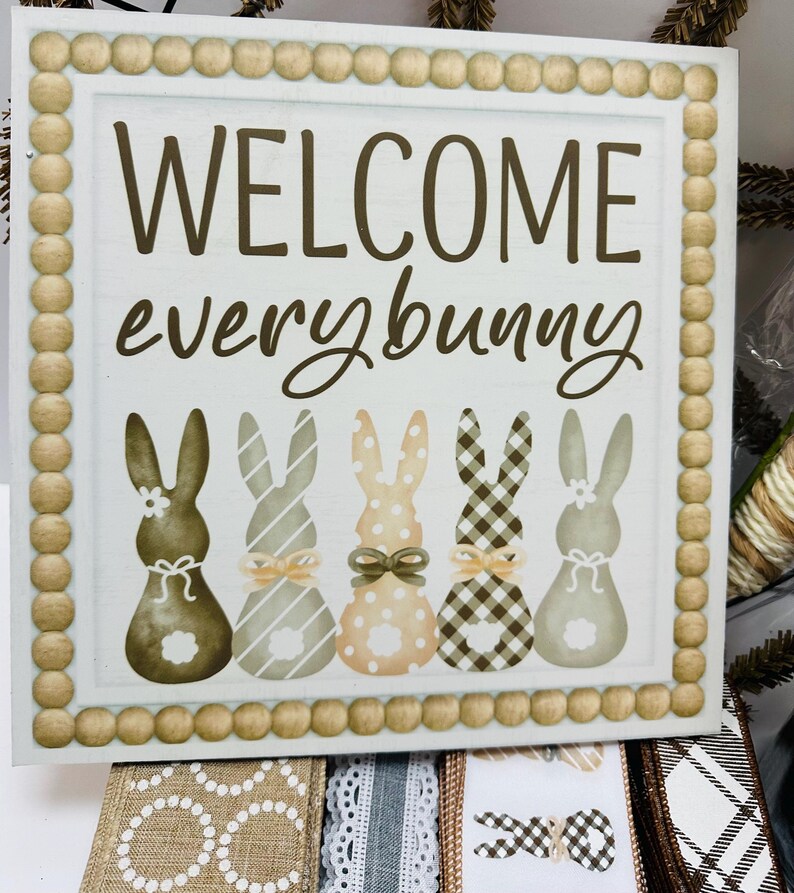 Peut inclure: Panneau carr&eacute; en bois avec l'inscription "WELCOME everybunny" en lettres marron. Le panneau pr&eacute;sente cinq silhouettes de lapins avec diff&eacute;rents motifs et n&oelig;uds. La bordure est faite de petites perles de bois rondes.
